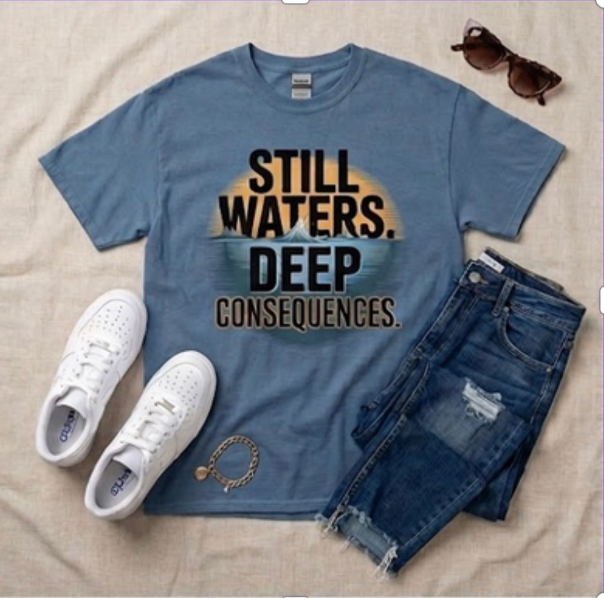 Blue t-shirt with 'Still Waters. Deep Consequences.' text, white sneakers, sunglasses, and chain on a beige surface.